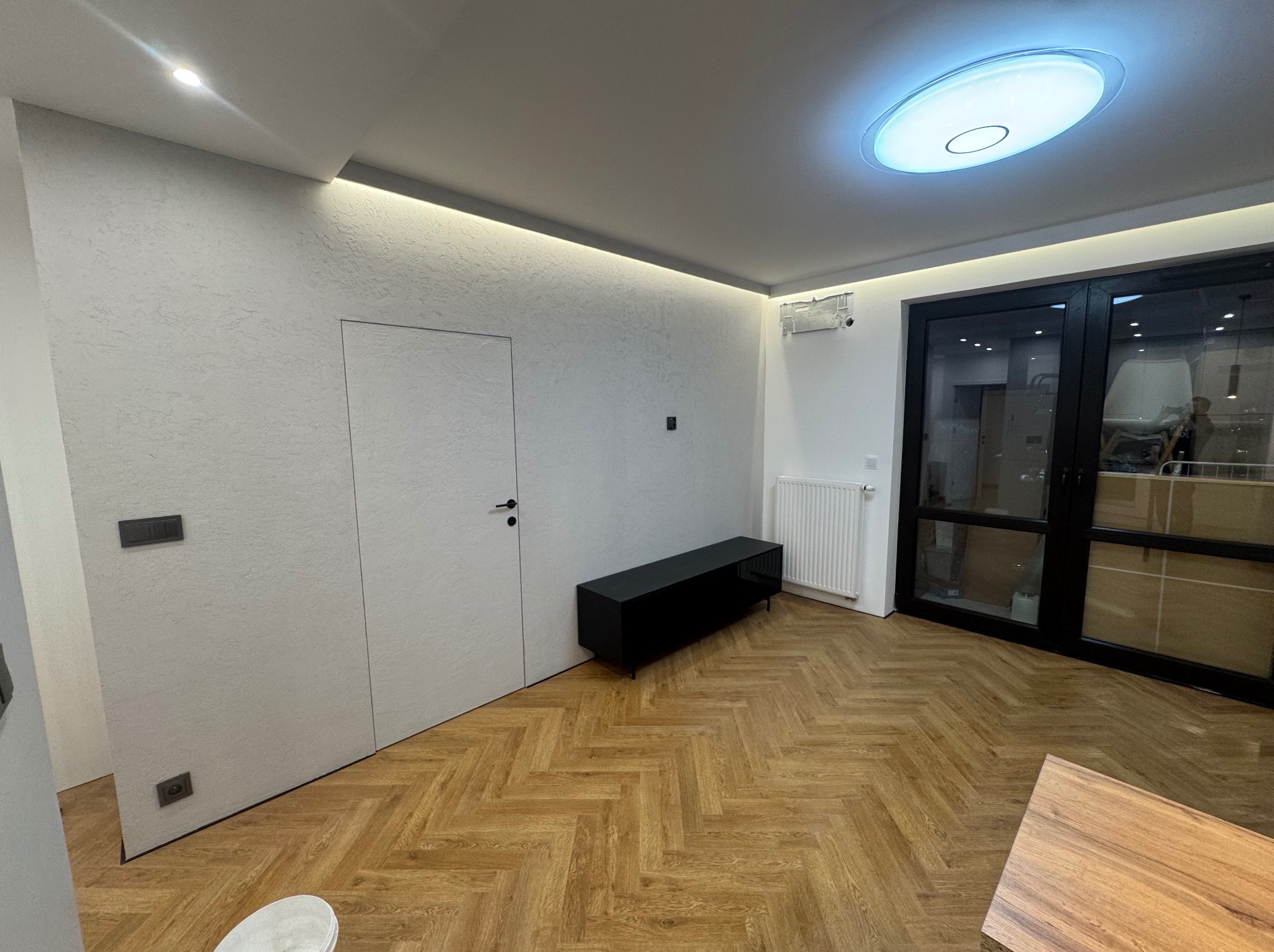 Salon po remoncie z parkietem ułożonym w jodełkę, oświetleniem LED w suficie i ukrytymi drzwiami w ścianie o fakturze baranka.