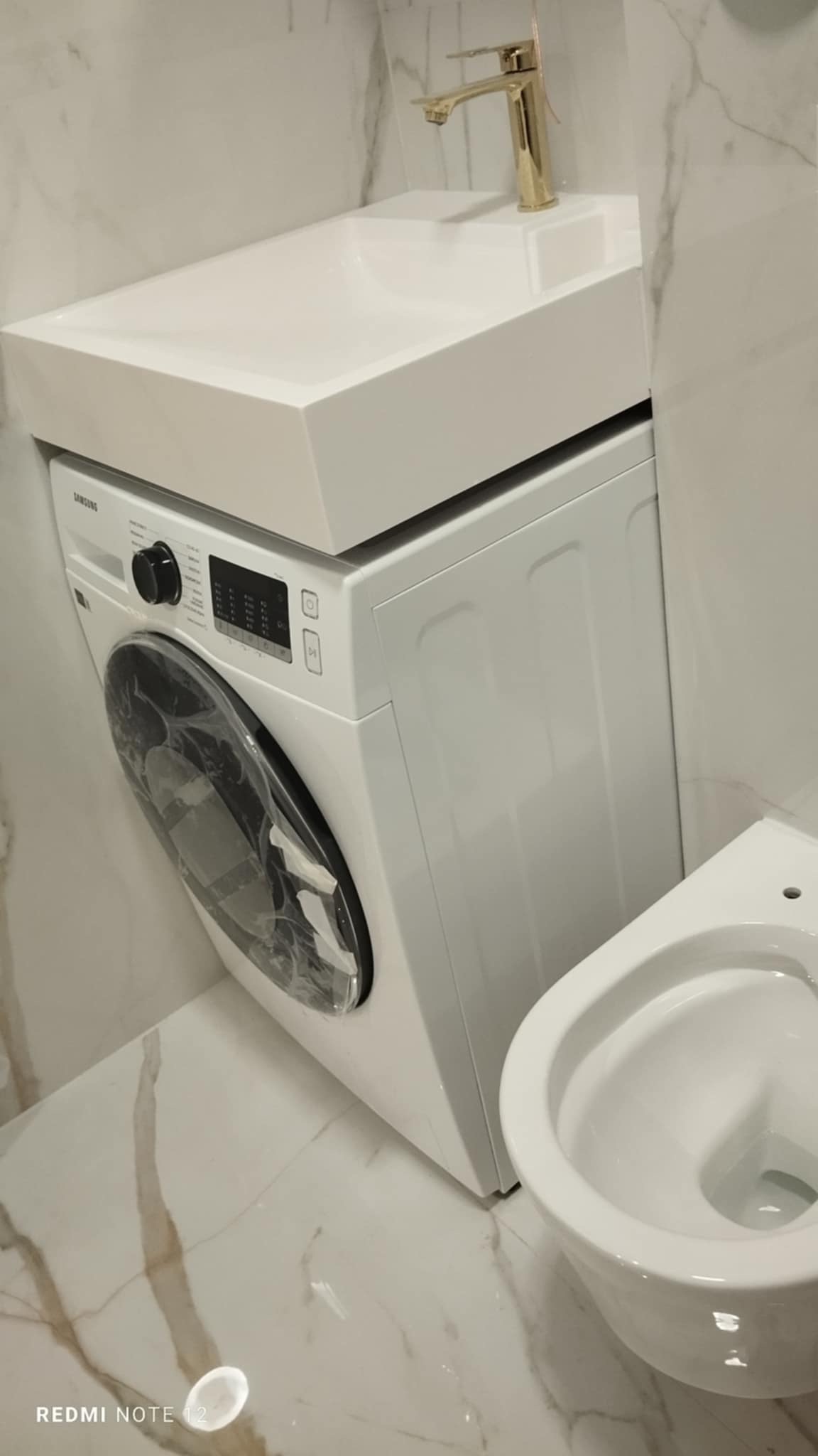 Kompaktowa łazienka z pralką Samsung pod umywalką nablatową i złotą armaturą, marmurowe płytki na ścianach i podłodze, obok toaleta.