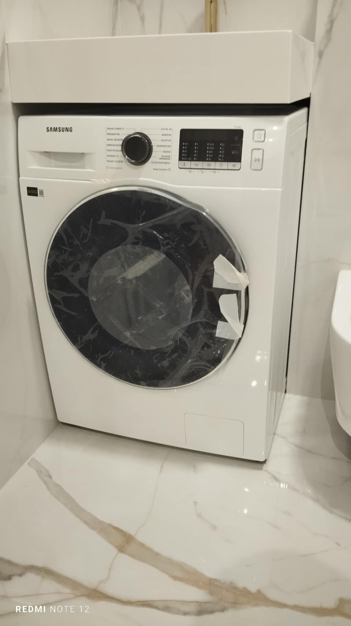Nowa biała pralka Samsung z folią ochronną na drzwiczkach, zamontowana w łazience z marmurową podłogą.