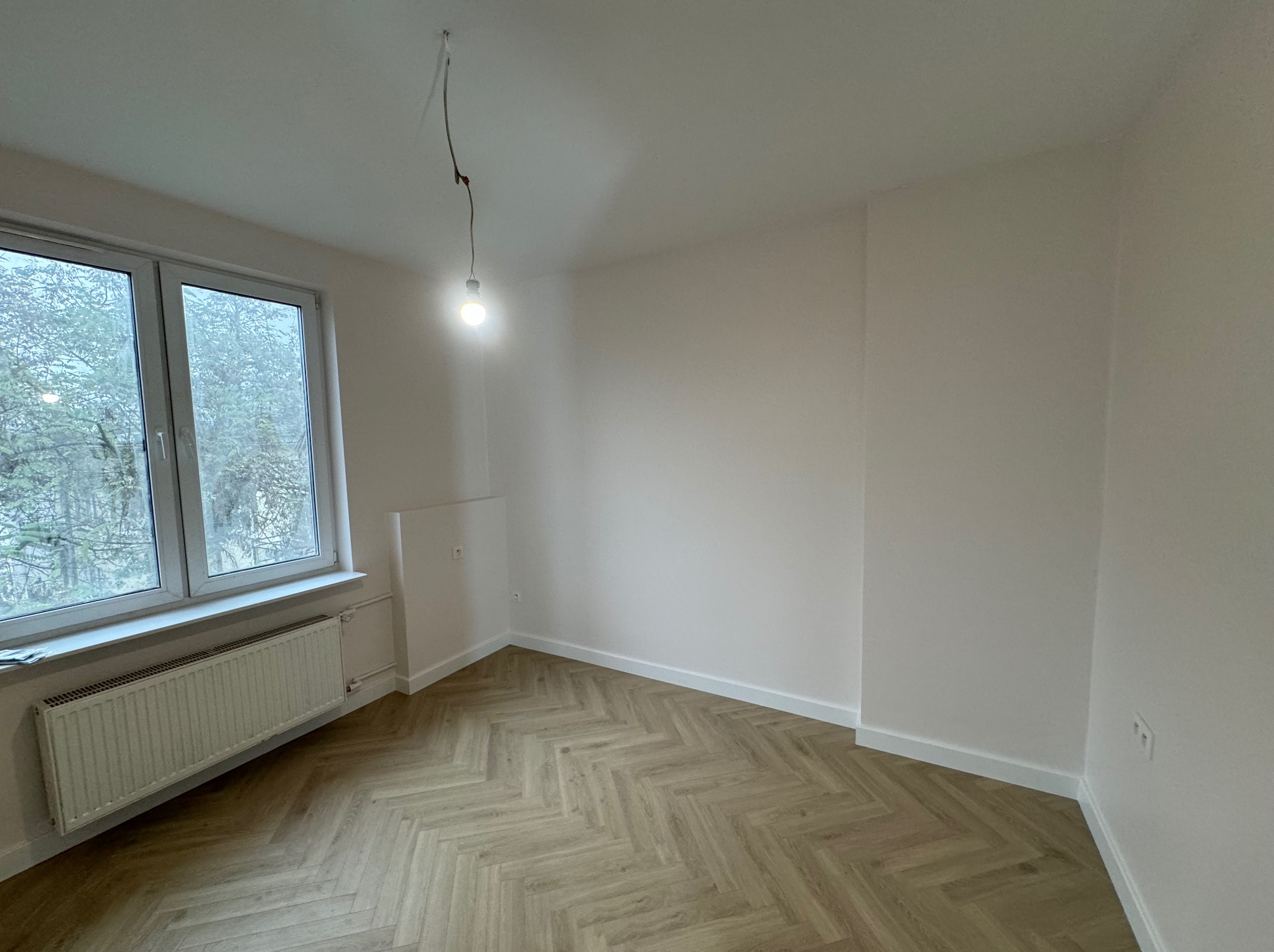 Pusty, odświeżony pokój z parkietem ułożonym w jodełkę, świeżo pomalowanymi ścianami na biało, grzejnikiem pod oknem i żarówką zwisającą z sufitu na kablu.