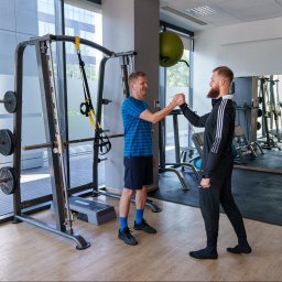 Trener personalny Kraków - Marcin Fiedorczyk - Trenuj Z Głową - Trening personalny Kraków - Trener personalny i klient w sportowym stroju witają się uściskiem dłoni w nowoczesnej siłowni z dużymi oknami i sprzętem do ćwiczeń.