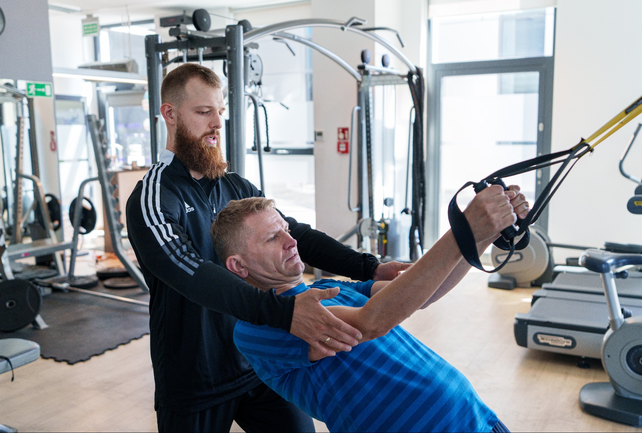 Mężczyzna w niebieskiej koszulce ćwiczy na maszynie z żółtymi linami pod okiem brodatego mężczyzny w czarnej bluzie z trzema białymi paskami na ramionach, w tle siłownia z różnymi maszynami.
