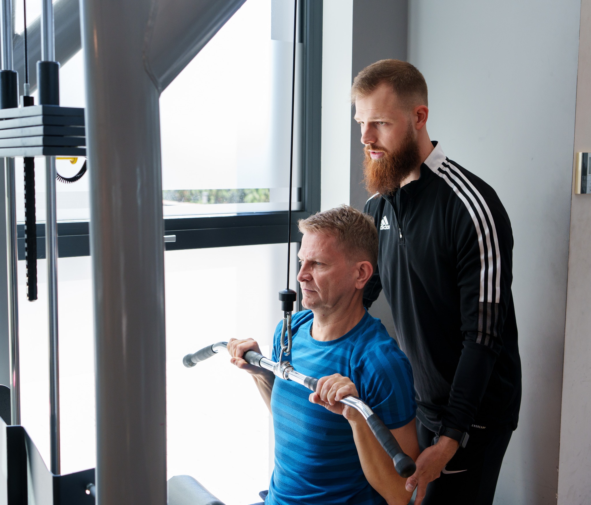 Mężczyzna ćwiczy na maszynie do wyciągania drążka pod nadzorem brodatego instruktora w czarnym dresie Adidas.