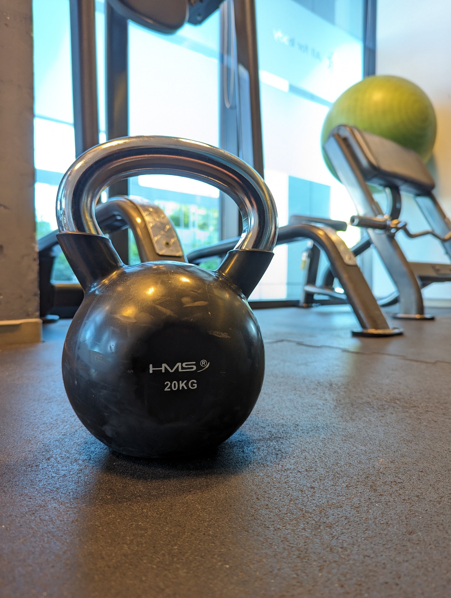 Czarny kettlebell 20kg marki HMS na tle sprzętu do ćwiczeń w siłowni, widok z niskiej perspektywy.