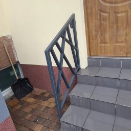 Antex Damian Brzozowski - Szara, metalowa balustrada o geometrycznym wzorze, zamontowana przy wejściu do domu z drewnianymi drzwiami i schodami wyłożonymi szarymi płytkami. Obok balustrady stoi czarna łopata do śniegu.