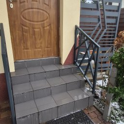 Antex Damian Brzozowski - Zewnętrzne schody wejściowe wyłożone szarą płytką, z metalową balustradą o geometrycznym wzorze, prowadzące do brązowych drzwi wejściowych.
