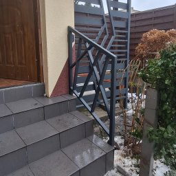 Antex Damian Brzozowski - Szare metalowe schody zewnętrzne z geometryczną balustradą, widok na drzwi wejściowe i fragment ogrodzenia w tle, pochmurny dzień.