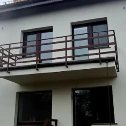 Balkon z brązową metalową balustradą na tle jasnej elewacji budynku z widocznymi oknami.