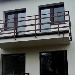 Antex Damian Brzozowski - Balkon z brązową, metalową balustradą na tle białej elewacji budynku z dwoma oknami na górze i dwoma na dole.