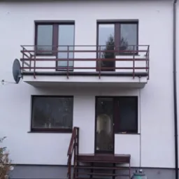 Biała elewacja domu z brązowymi oknami, balkonem z brązową balustradą i schodami wejściowymi, widoczna antena satelitarna i łańcuch spustowy.