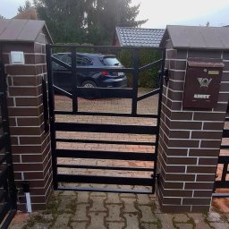 Antex Damian Brzozowski - Czarna brama wjazdowa z poziomymi szczeblami, zamontowana między dwoma murowanymi słupkami z brązowej cegły klinkierowej, na jednym ze słupków skrzynka pocztowa w stylu retro, na podjeździe kostka...