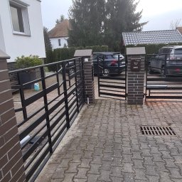 Antex Damian Brzozowski - Nowoczesna brama wjazdowa, czarna, metalowa, z elementami murowanymi z cegły, na posesji z kostki brukowej, z widocznymi samochodami w tle.