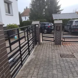 Nowoczesna brama wjazdowa, czarna, metalowa, z elementami murowanymi z cegły, na posesji z kostki brukowej, z widocznymi samochodami w tle.