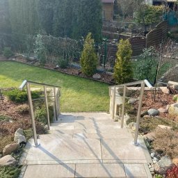 Antex Damian Brzozowski - Zewnętrzne schody z jasnych płyt z nierdzewnymi balustradami prowadzące do ogrodu z trawnikiem, tujami i kamieniami ozdobnymi.