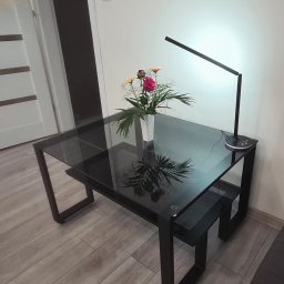 Antex Damian Brzozowski - Szklany stół z czarnymi nogami, na którym stoi wazon z kwiatami i nowoczesna lampka LED, oświetlająca kompozycję.