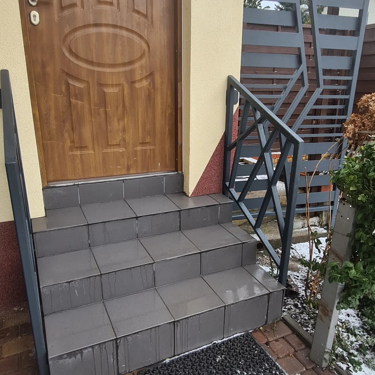 Zewnętrzne schody wejściowe wyłożone szarą płytką, z metalową balustradą o geometrycznym wzorze, prowadzące do brązowych drzwi wejściowych.
