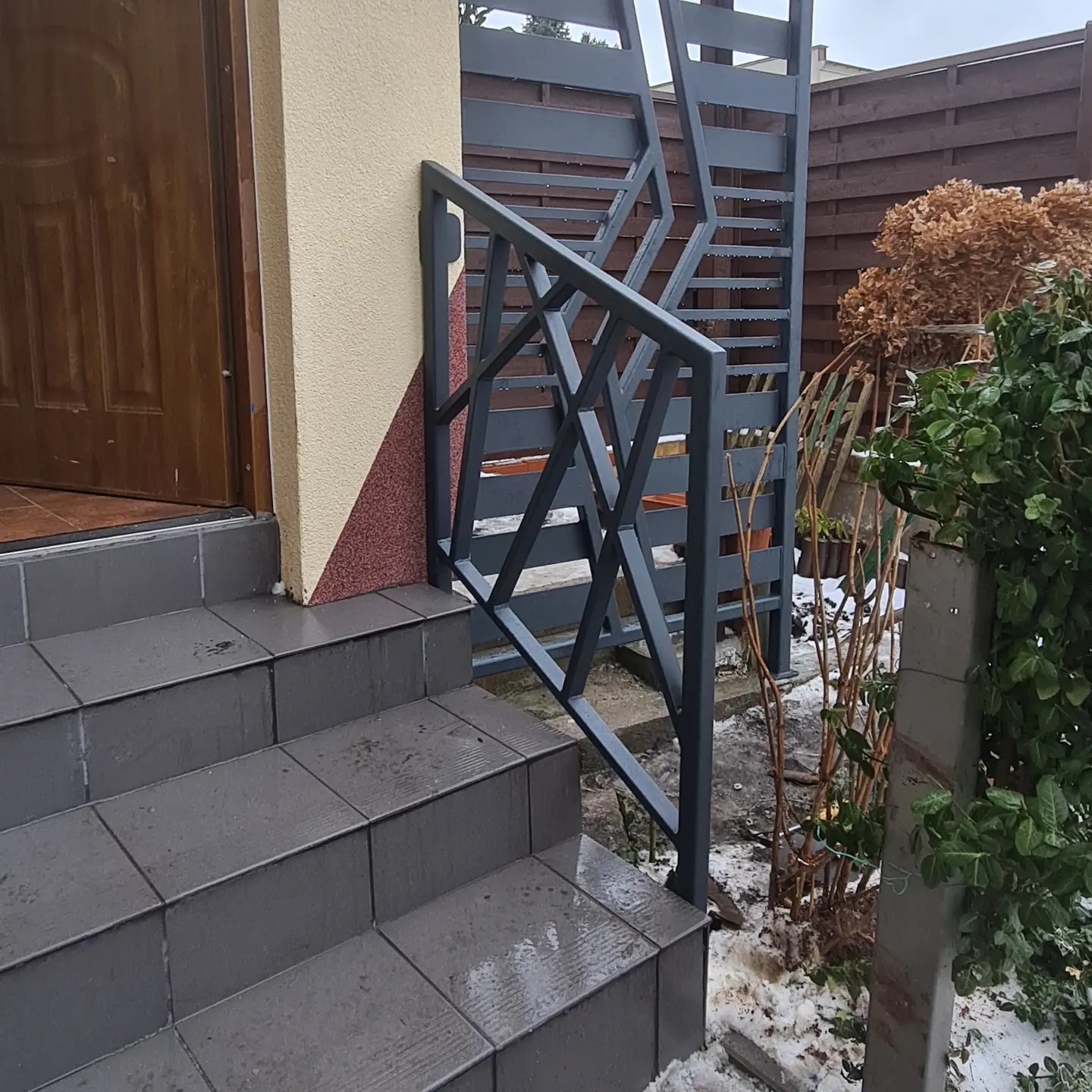 Szare metalowe schody zewnętrzne z geometryczną balustradą, widok na drzwi wejściowe i fragment ogrodzenia w tle, pochmurny dzień.