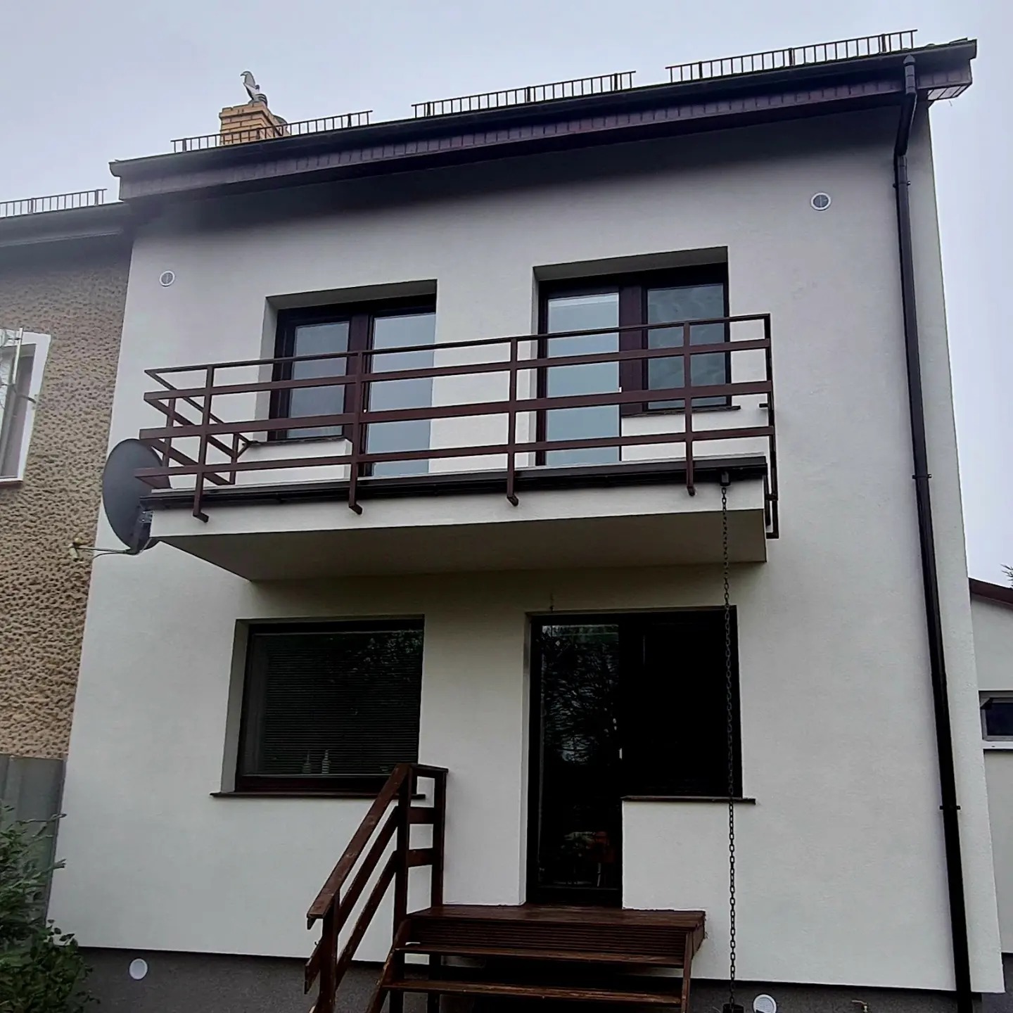 Dwupiętrowy dom po renowacji elewacji, z brązowymi oknami, balkonem z balustradą i schodami wejściowymi; na kominie siedzi ptak.