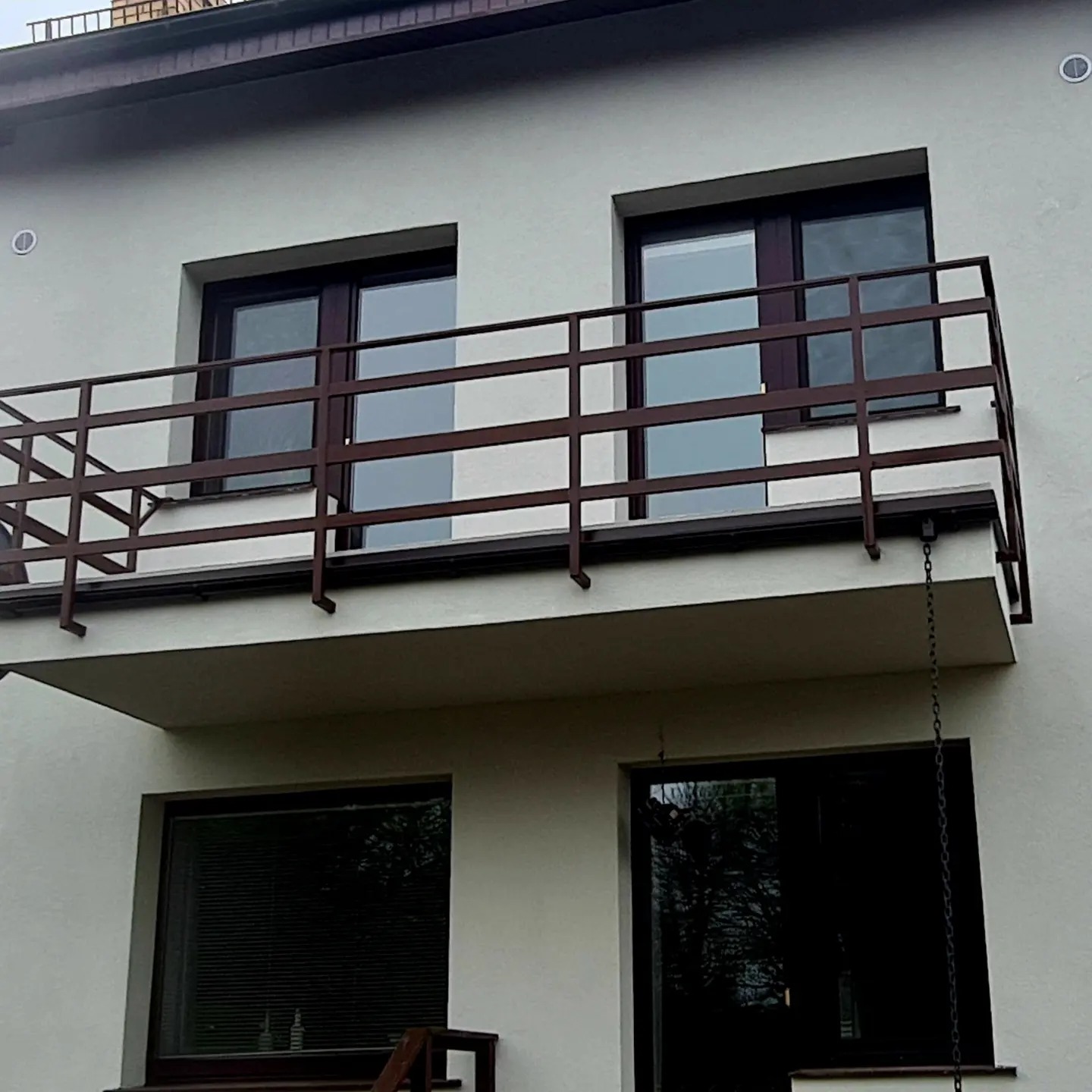 Balkon z brązową metalową balustradą na tle jasnej elewacji budynku z widocznymi oknami.