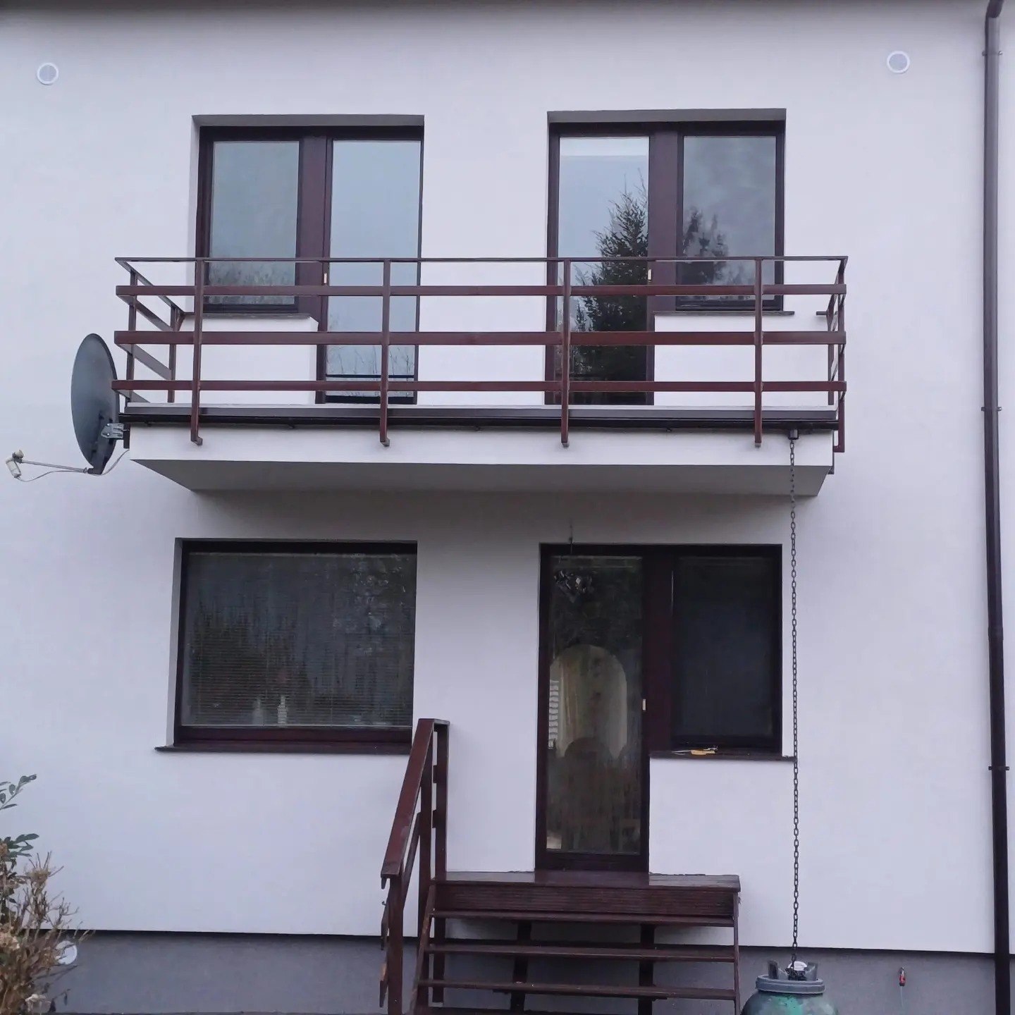 Biała elewacja domu z brązowymi oknami, balkonem z brązową balustradą i schodami wejściowymi, widoczna antena satelitarna i łańcuch spustowy.