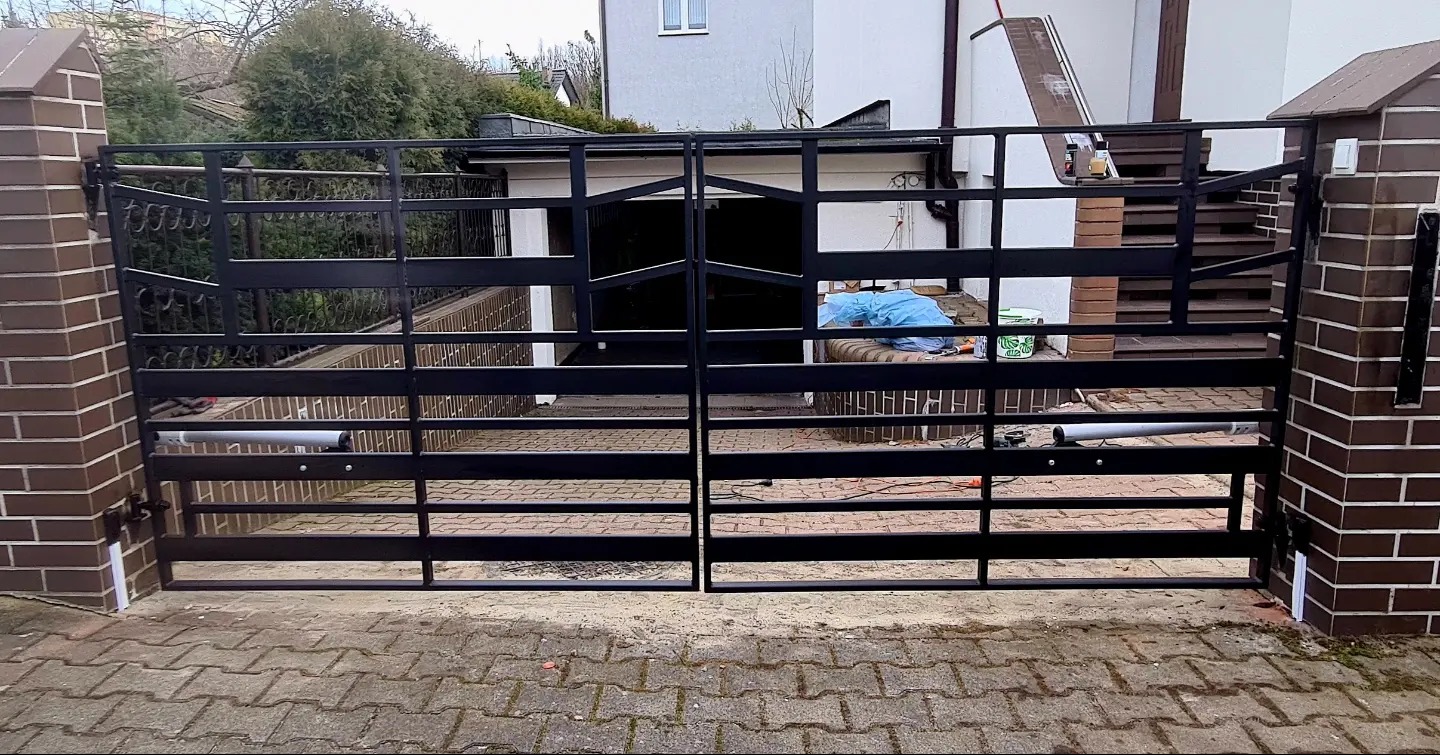 Czarna, metalowa brama wjazdowa dwuskrzydłowa z automatyką, zamontowana na ceglanych słupkach, widok z zewnątrz posesji.