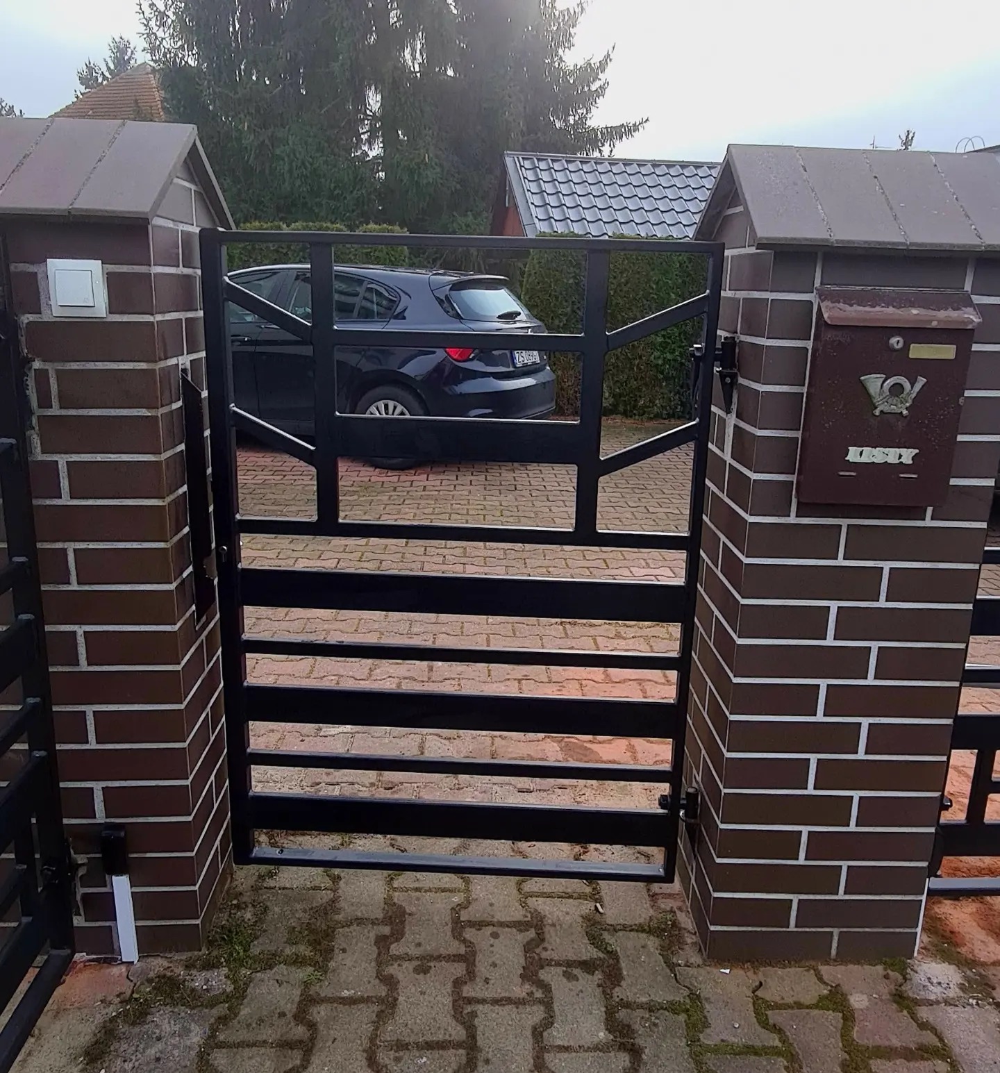 Czarna brama wjazdowa z poziomymi szczeblami, zamontowana między dwoma murowanymi słupkami z brązowej cegły klinkierowej, na jednym ze słupków skrzynka pocztowa w stylu retro, na podjeździe kostka...