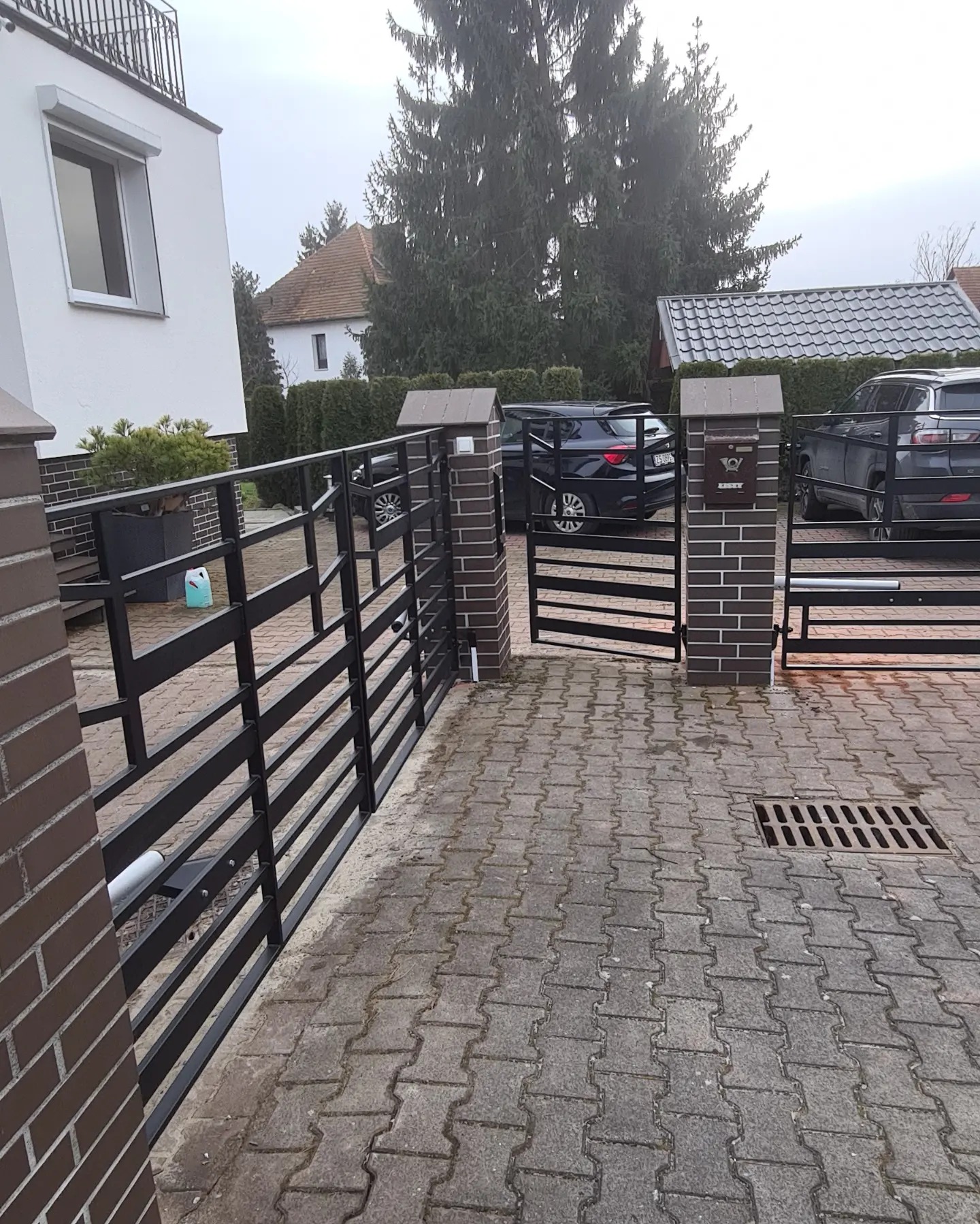 Nowoczesna brama wjazdowa, czarna, metalowa, z elementami murowanymi z cegły, na posesji z kostki brukowej, z widocznymi samochodami w tle.