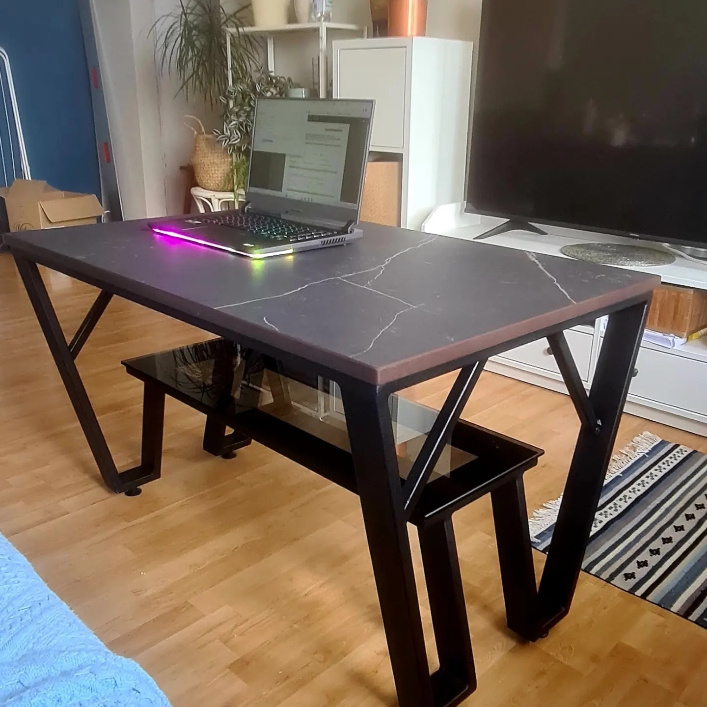 Czarny metalowy stół z blatem imitującym marmur w nowoczesnym salonie, z laptopem z podświetlaną klawiaturą, na drewnianej podłodze, obok wzorzysty dywanik.
