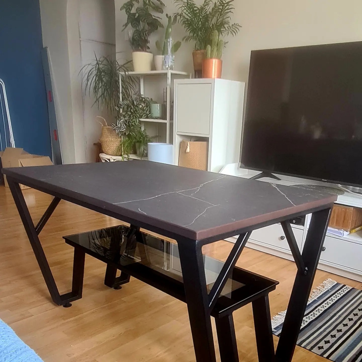 Stół z blatem imitującym marmur, czarne metalowe nogi o geometrycznym kształcie, dodatkowa szklana półka, w tle rośliny doniczkowe i telewizor.