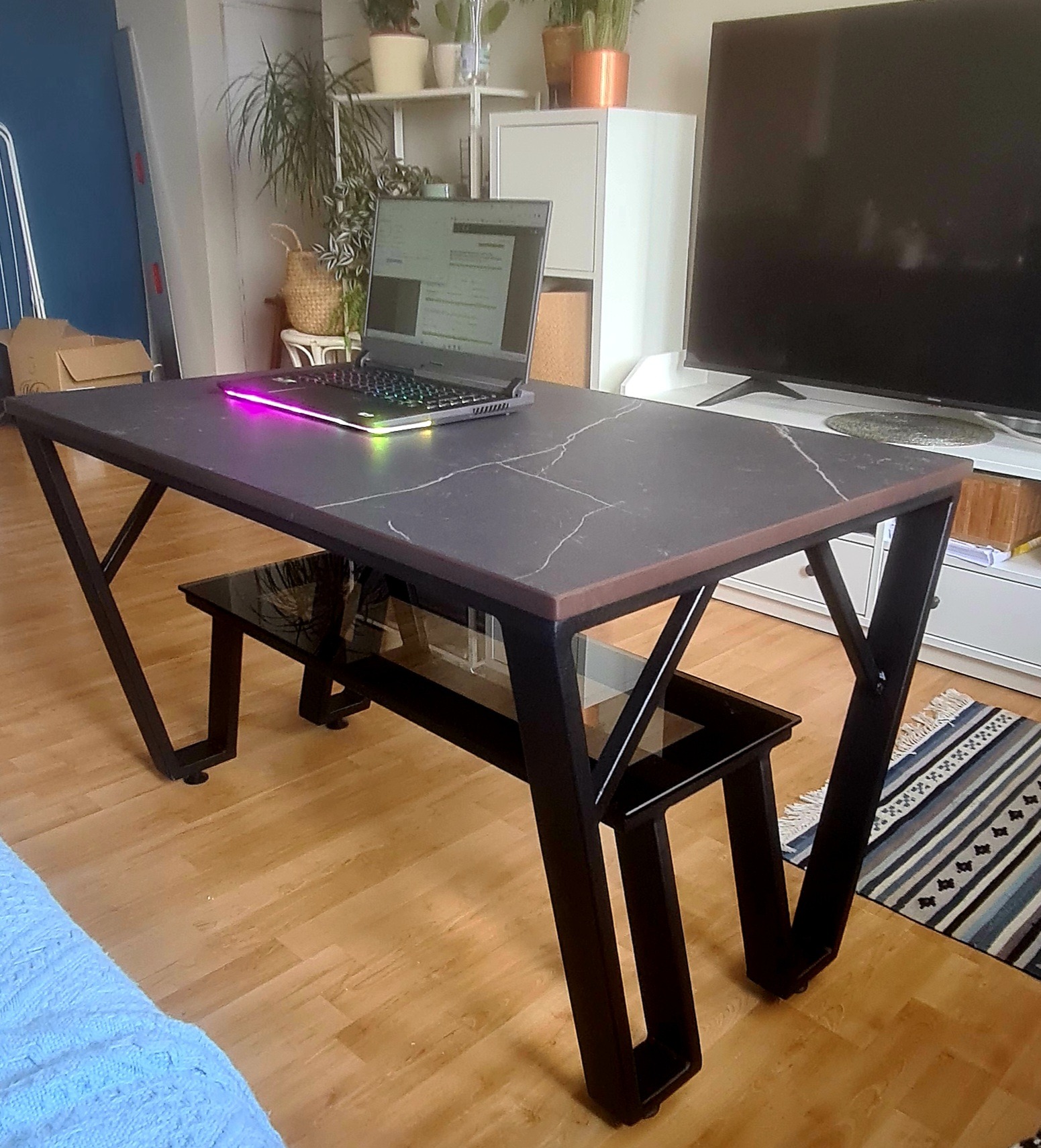 Czarny biurko z blatem imitującym marmur i podświetlanym laptopem, nowoczesny design z geometrycznymi nogami i szklaną półką.