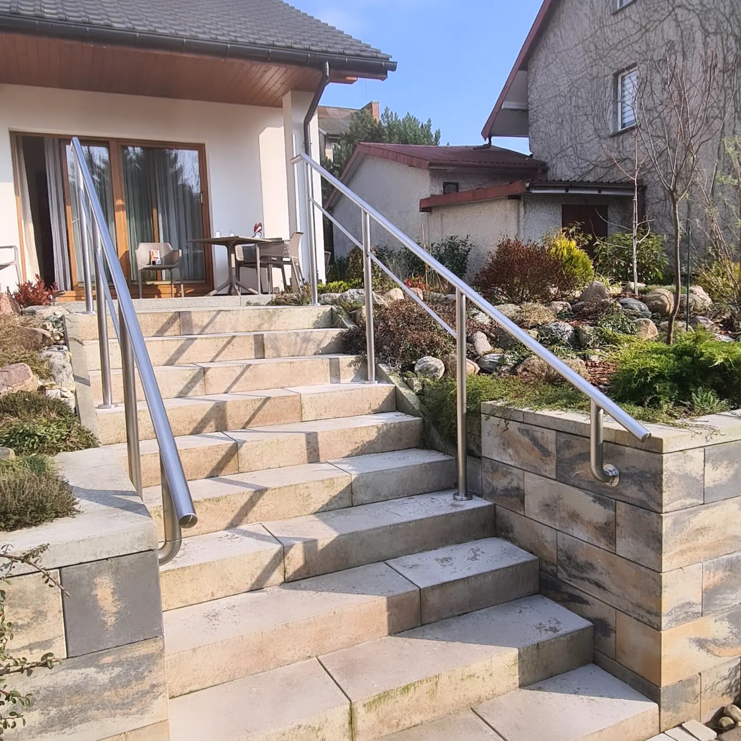 Zewnętrzne schody z jasnych, blokowych stopni prowadzące do domu, zabezpieczone nowoczesną balustradą ze stali nierdzewnej, w tle zadbany ogród i budynek pokryty bluszczem.