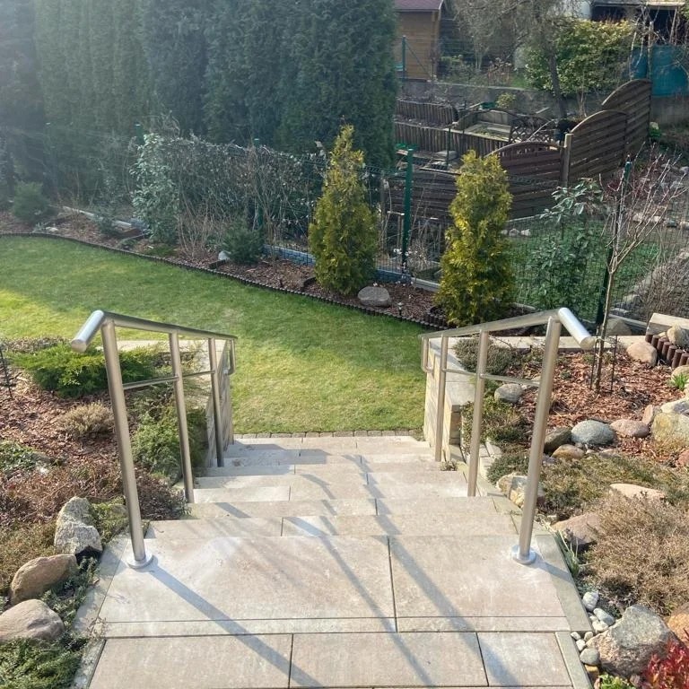 Zewnętrzne schody z jasnych płyt z nierdzewnymi balustradami prowadzące do ogrodu z trawnikiem, tujami i kamieniami ozdobnymi.