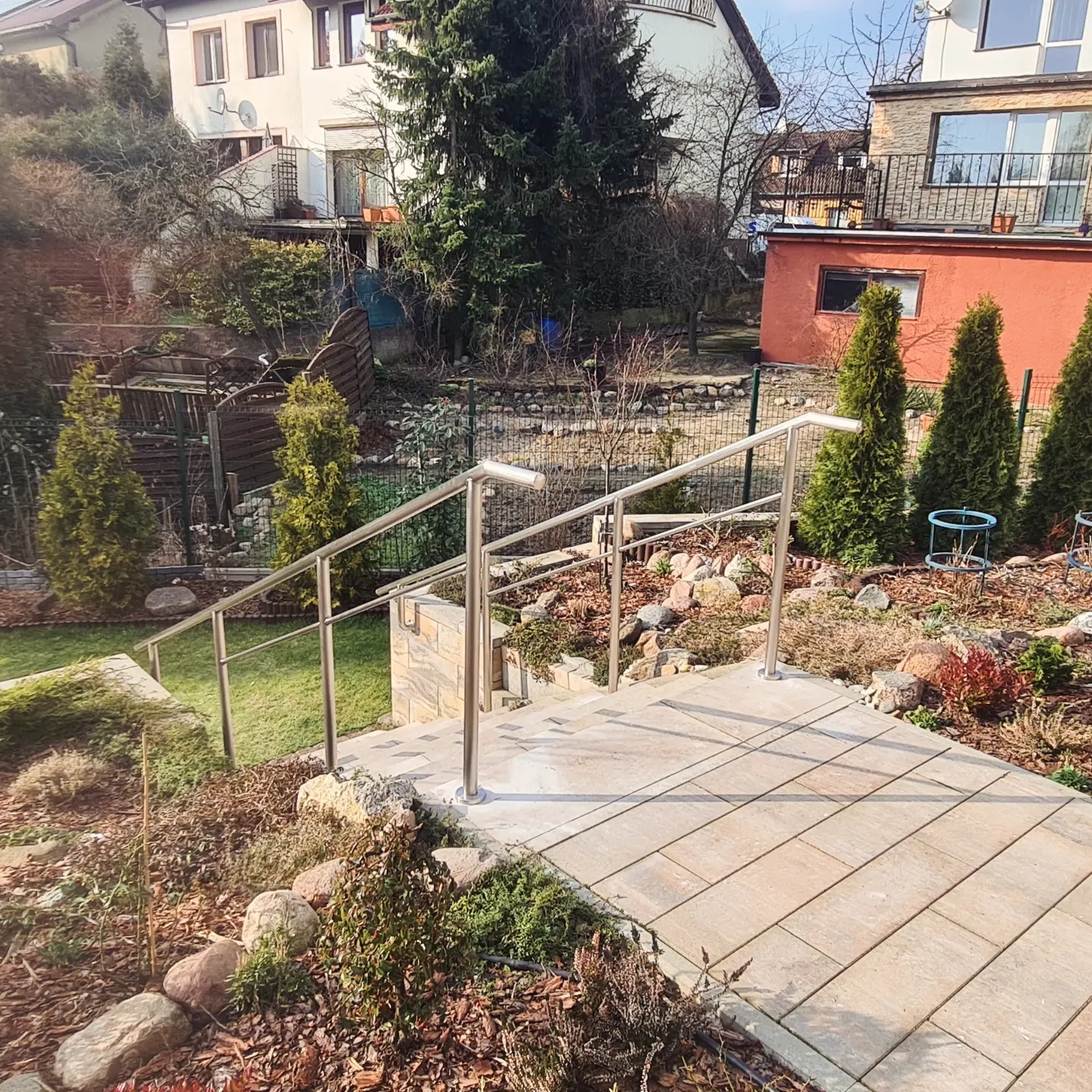 Zewnętrzne schody wyłożone jasną kostką brukową z balustradą ze stali nierdzewnej, prowadzące w dół ogrodu z widokiem na trawnik i roślinność ozdobną.