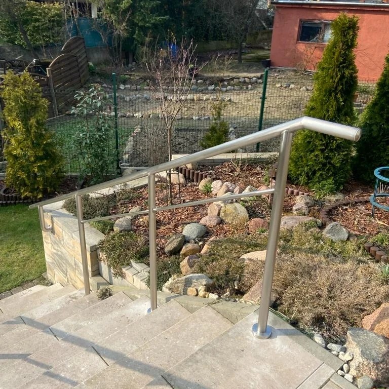 Zewnętrzne schody z jasnych płyt z zamontowaną, prostą balustradą ze stali nierdzewnej w Police, otoczone zielenią i kamieniami.