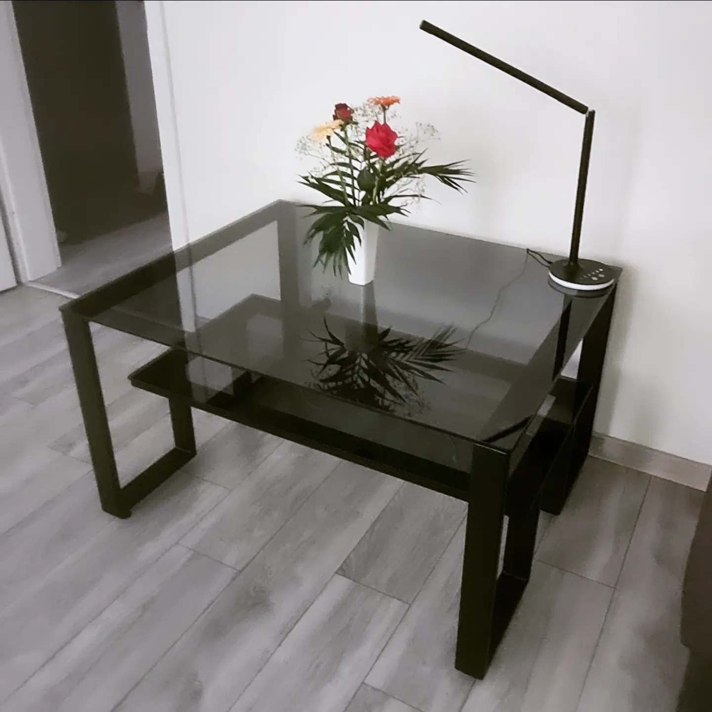 Nowoczesny stolik kawowy z czarną ramą i szklanym blatem, na którym stoi wazon z kwiatami i lampka biurkowa w minimalistycznym stylu.