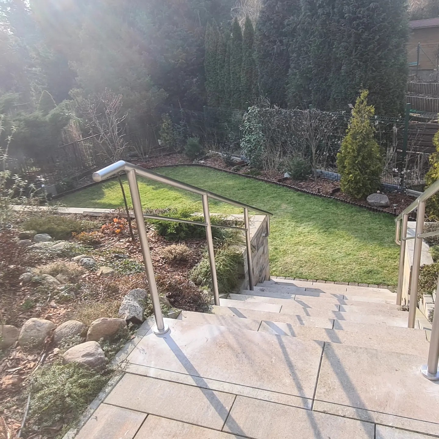 Zewnętrzne schody z jasnych płyt z nierdzewną balustradą prowadzące do ogrodu z trawnikiem i ozdobnymi roślinami, oświetlone silnym słońcem.