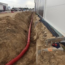 Usługi Elektryczne Marek Kozieł - Czerwony karbowany przewód ułożony w wykopie wzdłuż fundamentu budynku przemysłowego. Widoczne betonowe elementy fundamentu, ziemia i w oddali sylwetka pracownika.
