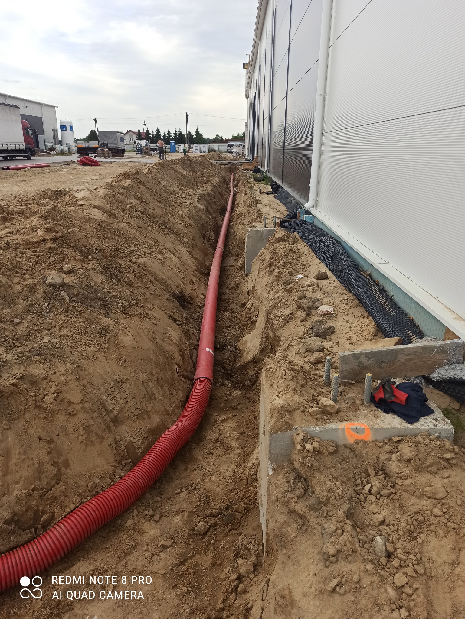 Czerwony karbowany przewód ułożony w wykopie wzdłuż fundamentu budynku przemysłowego. Widoczne betonowe elementy fundamentu, ziemia i w oddali sylwetka pracownika.