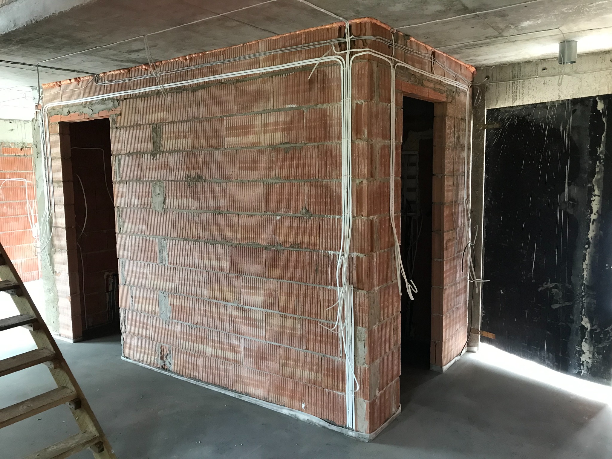 Wewnętrzne ściany z cegły dziurawki w trakcie budowy, z widocznymi przewodami elektrycznymi prowadzonymi po wierzchu i zabezpieczonymi pianką montażową pod sufitem, na surowym betonowym stropie.
