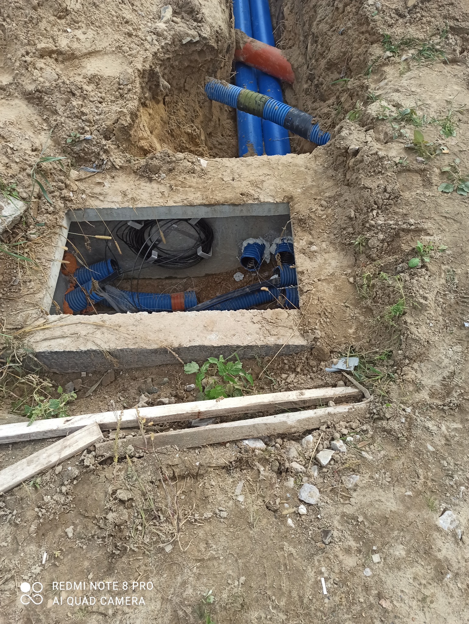 Wykop z betonową obudową, wewnątrz niebieskie rury karbowane i czarne przewody elektryczne, część rur wystaje z wykopu w ziemi.