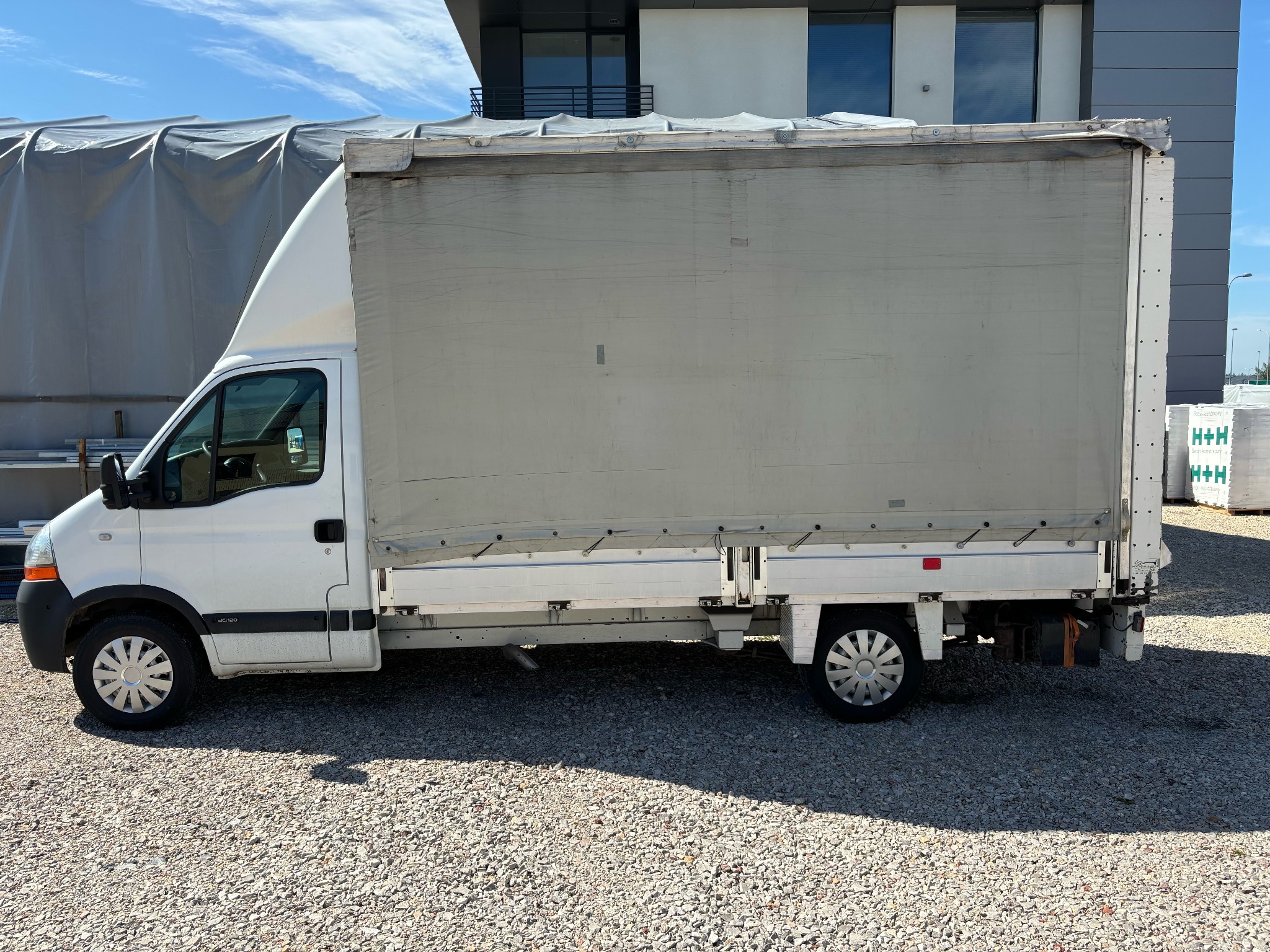 Biały samochód dostawczy z plandeką na żwirowym podłożu, widok z boku, z paletami cegieł w tle.