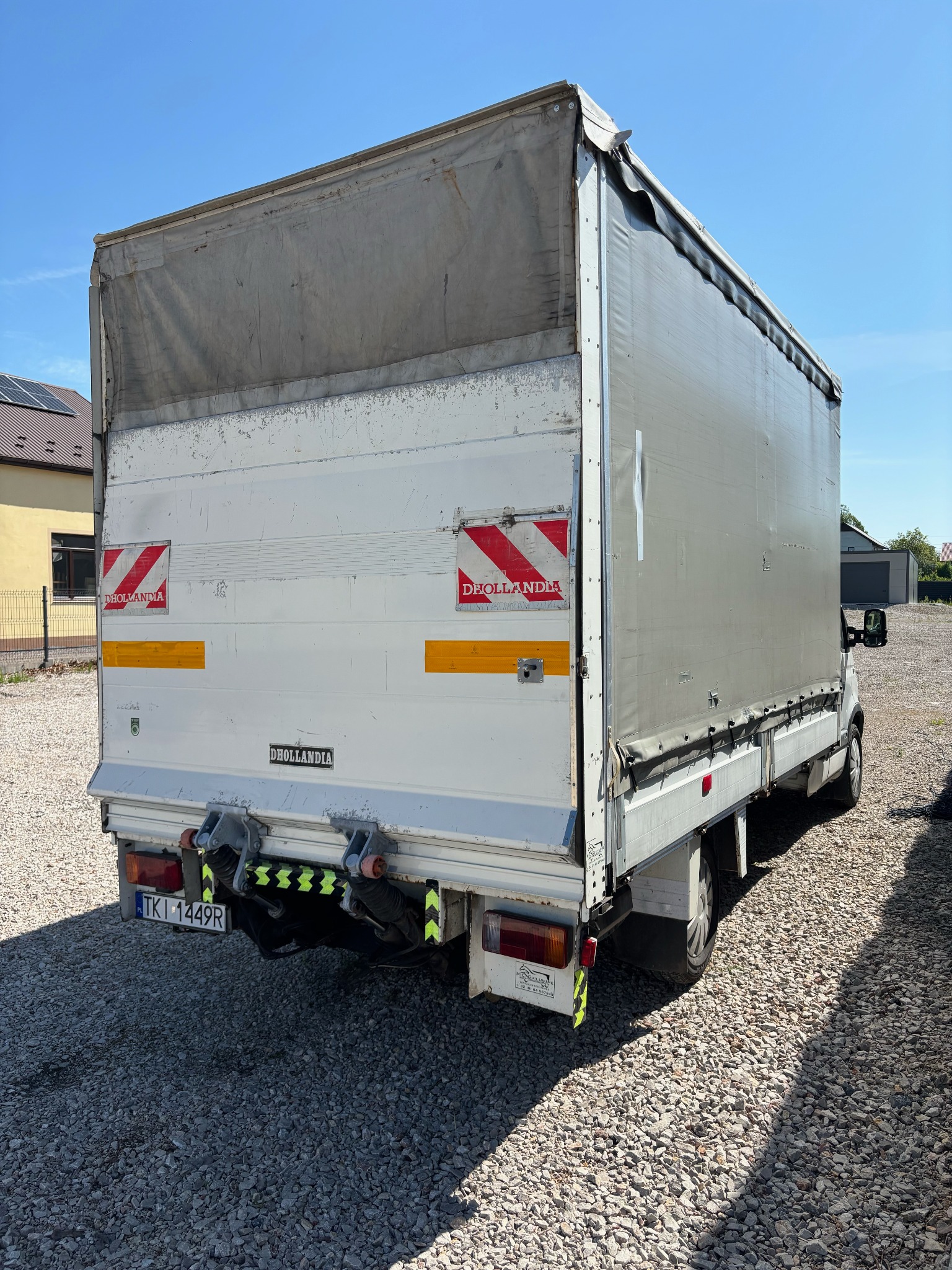 Tył białego samochodu dostawczego z plandeką, widoczne oznaczenia ostrzegawcze, tablica rejestracyjna i podnośnik hydrauliczny.