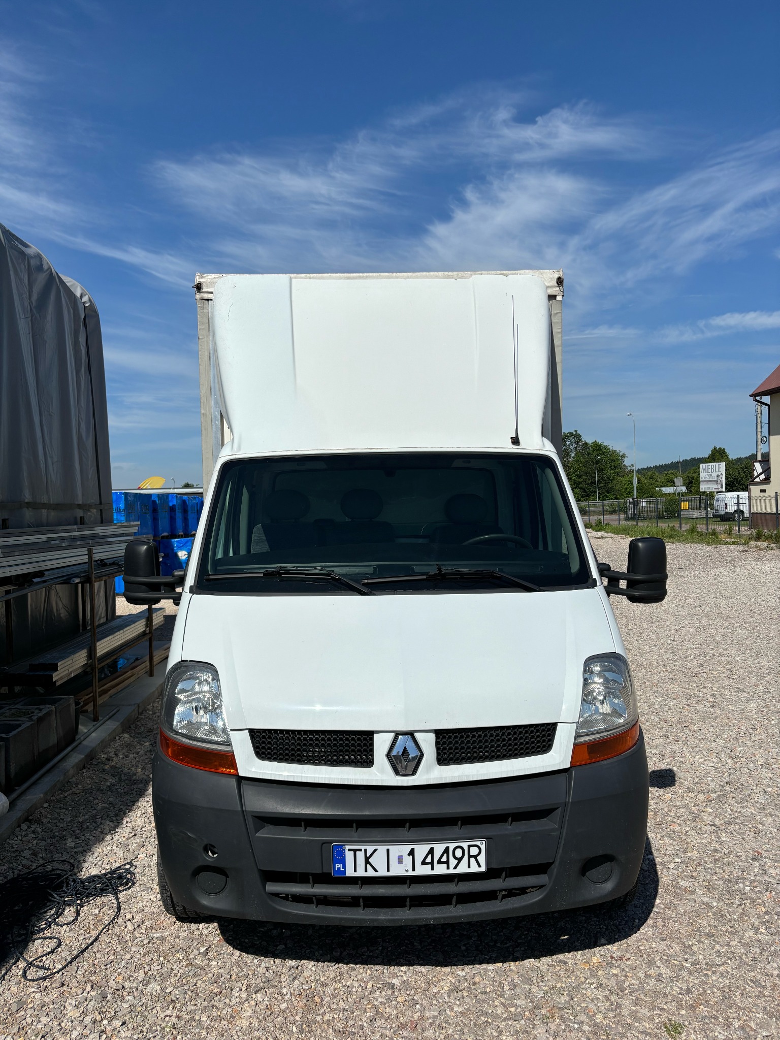 Biały samochód dostawczy Renault Master z wysoką zabudową, zaparkowany na żwirowym placu w słoneczny dzień, widok z przodu na tle błękitnego nieba z delikatnymi chmurami.