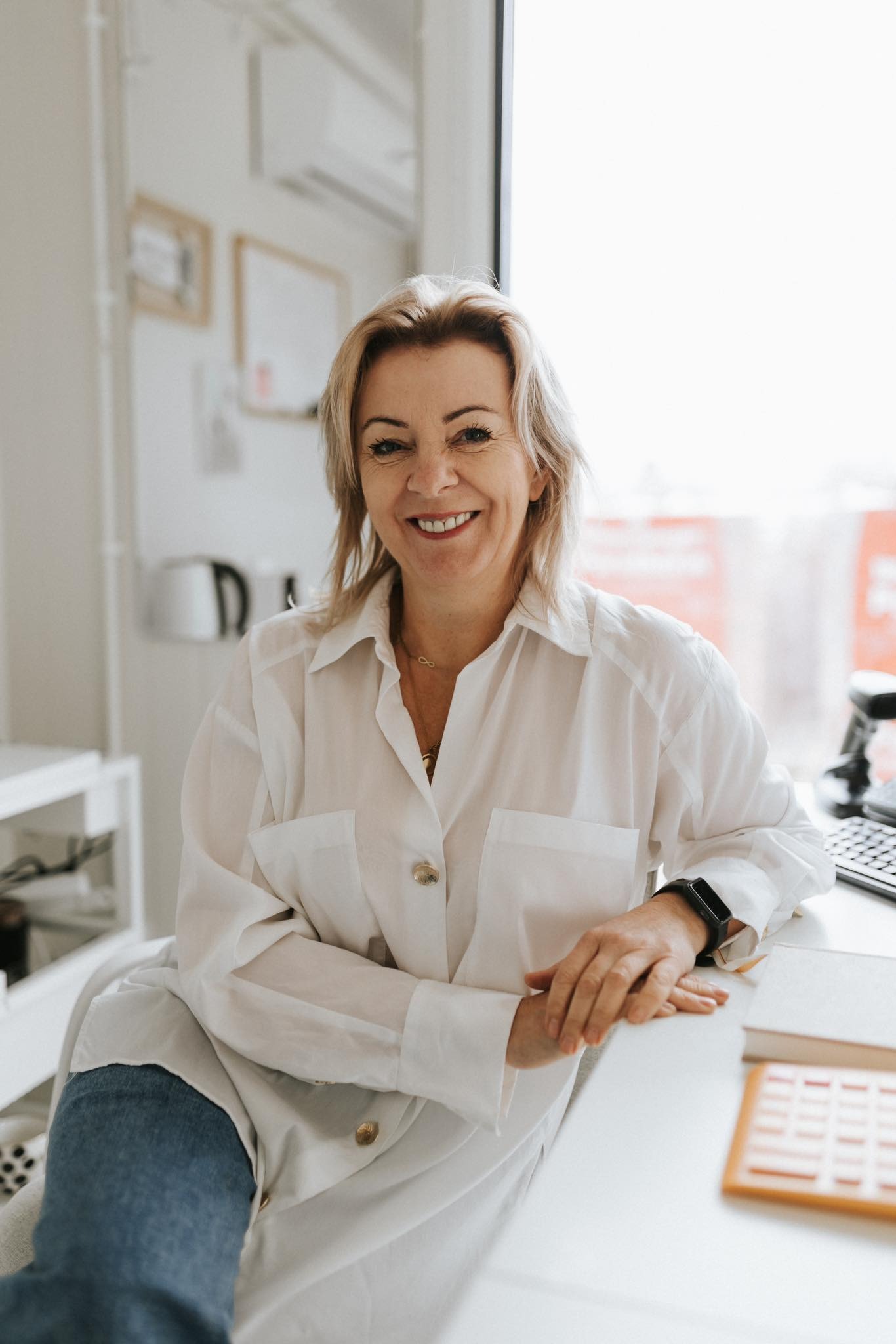Uśmiechnięta kobieta w białej koszuli siedzi przy biurku w jasnym pomieszczeniu, na biurku leżą notatniki i kalendarz.