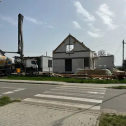 Wznoszenie ścian domu z betonu komórkowego w stanie surowym otwartym, widoczny betonowóz z pompą do betonu oraz składowane drewno budowlane.