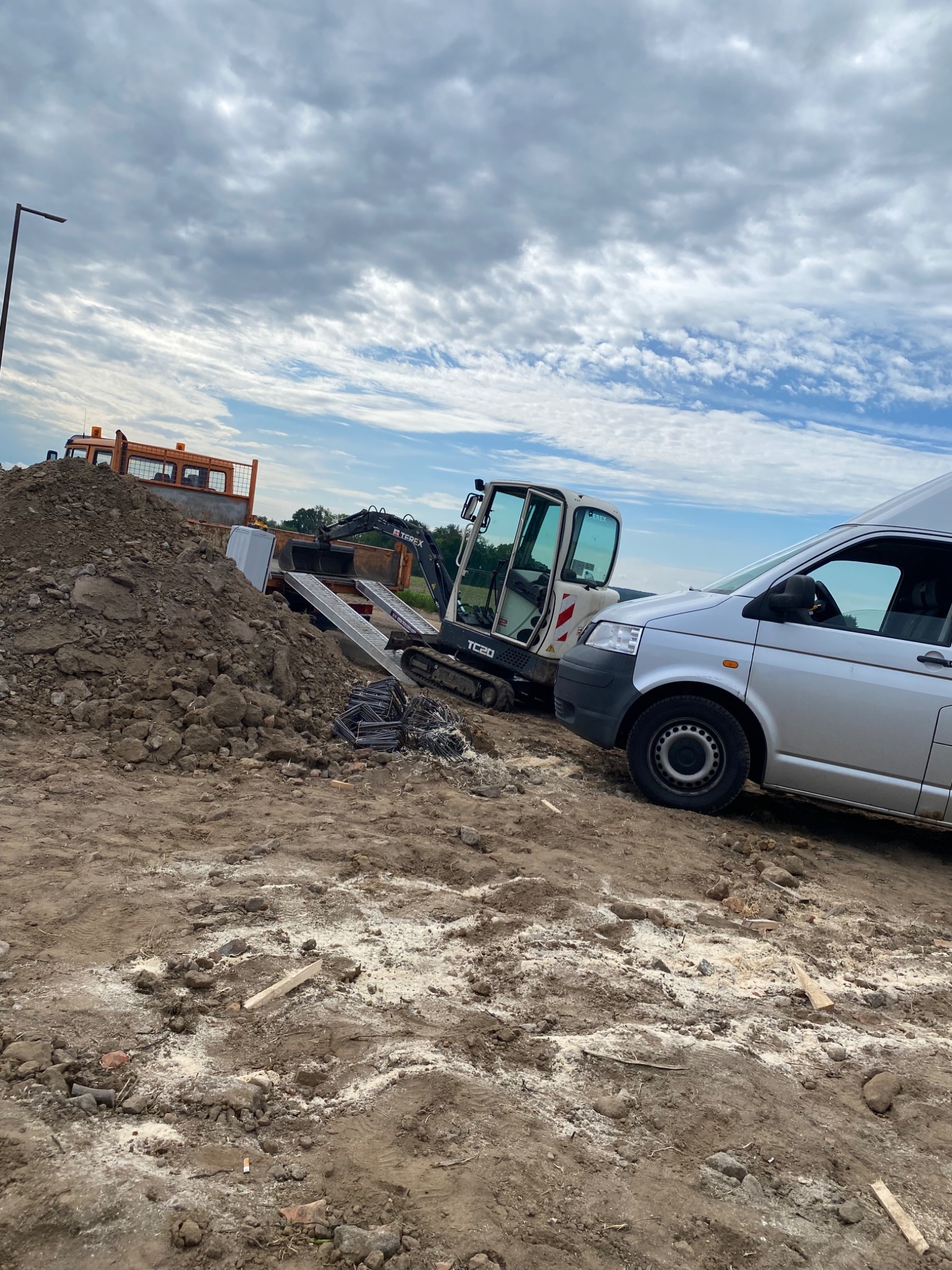 Mała koparka Terex TC20 z otwartą kabiną, zaparkowana obok srebrnego samochodu dostawczego na placu budowy z hałdą ziemi w tle, w pochmurny dzień.