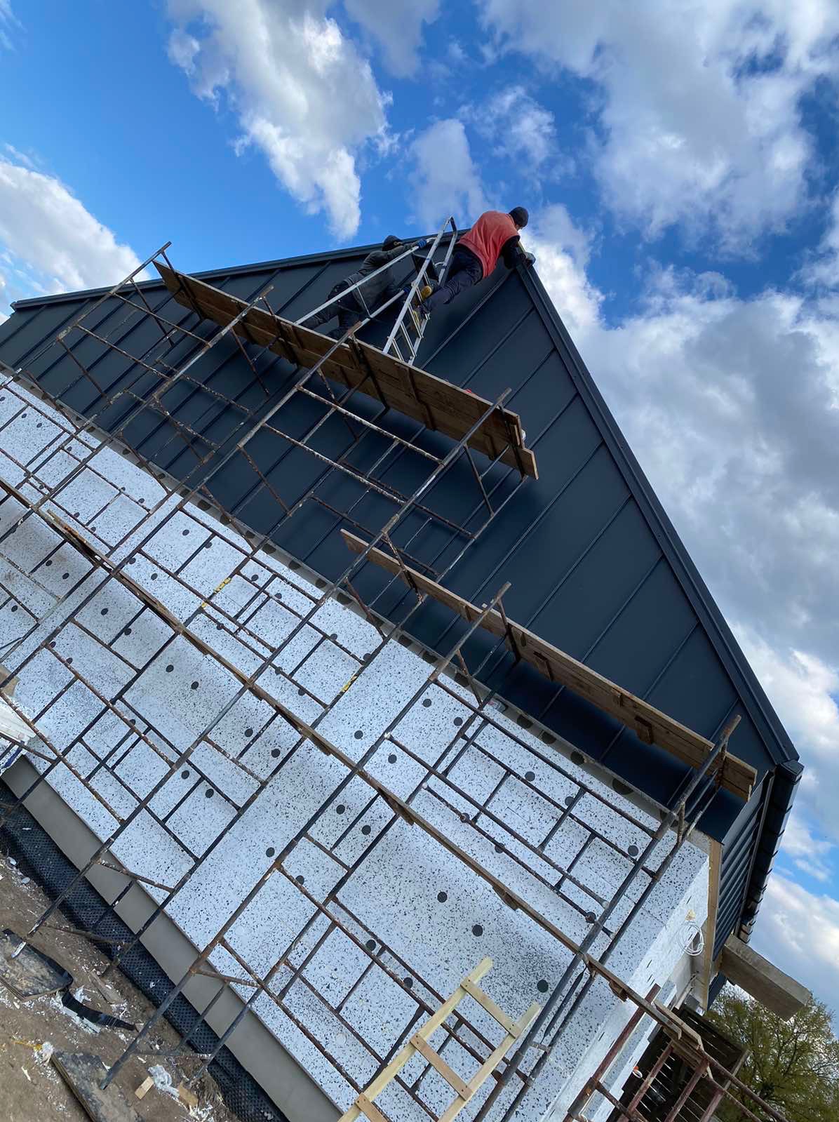 Ocieplanie elewacji budynku jednorodzinnego z użyciem rusztowania i widocznym fragmentem dachu z blachy na rąbek stojący, pracownik na drabinie montuje obróbki blacharskie.