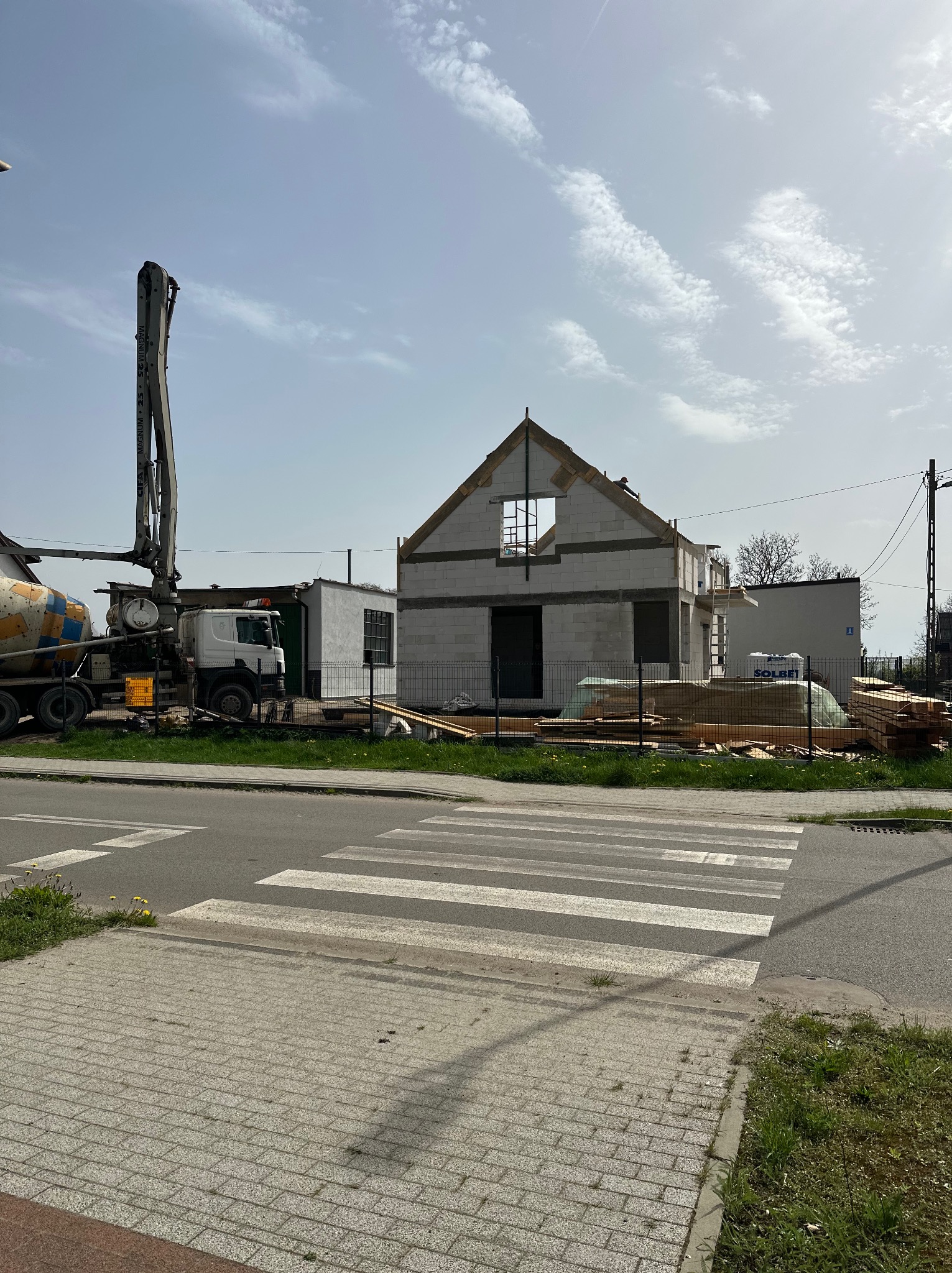 Wznoszenie ścian domu z betonu komórkowego w stanie surowym otwartym, widoczny betonowóz z pompą do betonu oraz składowane drewno budowlane.