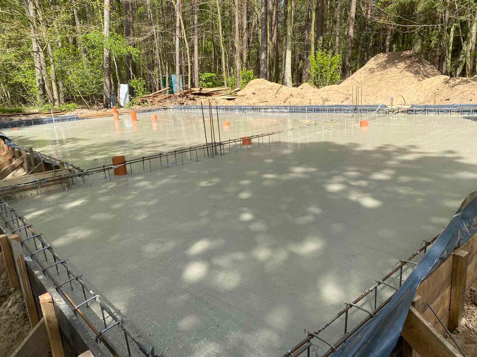 Wylany, mokry fundament pod budowę domu, z widocznym zbrojeniem i szalunkiem, na tle drzew.