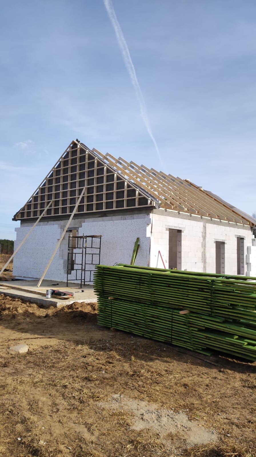 Wznoszony budynek jednorodzinny z białego bloczka, z widoczną konstrukcją dachu z drewnianych łat i kontrłat oraz czarną membraną dachową, obok leży stos zielonych impregnowanych desek.